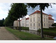 Schlossgebäude in klassizistischem Stil mit cremefarbenen Fassaden und roten Dächern, von einem Wassergraben umgeben. Im Vordergrund verläuft ein Kiesweg entlang einer Allee aus Bäumen.