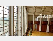 Großer Raum mit hohen Fenstern aus milchigem Glas in rechteckigen Segmenten, einer hölzernen Wandverkleidung und Deckengestaltung mit rechteckigen Ornamenten. Modernes Kronleuchtersystem mit kugelförmigen Lampen sowie Pflanzen und ein Metallständer im Vordergrund.