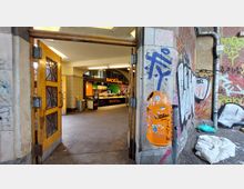 Offener Eingang eines Backwarenladens mit der Aufschrift „BACKBaron“ in einem urbanen Bereich. Außen sind eine orangefarbene Mülltonne und Wände mit Graffiti sichtbar; im Inneren des Ladens befinden sich eine beleuchtete Theke und geflieste Wände.