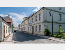 Kopfsteinpflasterstraße in einem historischen Stadtbereich, gesäumt von überwiegend hellen, zweigeschossigen Gebäuden mit Satteldächern und klassischen Fenstern. Im Vordergrund ein schmiedeeiserner Laternenpfahl, im Hintergrund Autos und weitere Häuser unter klarem Himmel.