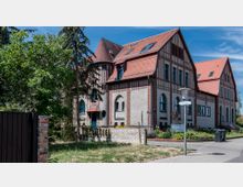 Historisches Gebäude mit roten Ziegeldächern und verputzten Wänden, durchsetzt mit roten Klinkerelementen, gotischen Fensterbögen und einem kleinen Vordach. Umgeben von Bäumen, einem Vorgarten sowie einem Gehweg mit einer Straßenlaterne und Straßenschildern.