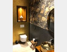 Modern gestaltetes Badezimmer mit goldener Akzentgestaltung, darunter eine gemusterte Tapete mit Flamingomotiven und einer Wand aus kleinen, goldfarbenen Kacheln. Zu sehen sind ein wandhängendes WC, ein goldglänzendes Waschbecken auf einem Holzmöbel sowie eine beleuchtete Nische mit einer Buddha-Statue.