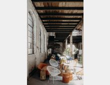 Ein Industrieraum mit unverputzten weißen Ziegelwänden, großen Bogenfenstern und einer offenen Holzbalkendecke. Der Raum ist mit modernen, farbigen Möbeln dekoriert, darunter orangefarbene und blaue Sessel sowie weiße Stühle, und im Hintergrund führt eine Metalltreppe zum oberen Bereich.