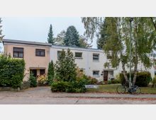 Eine Reihe flacher Reihenhäuser mit pastellfarbenen Fassaden, umgeben von kleinen Vorgärten mit Bäumen und Sträuchern. Im Vordergrund stehen ein Fahrrad an einer Straßenlaterne und ein Straßenschild mit Hausnummern.