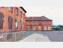 Backstein-Gebäude mit mehreren zugemauerten Fenstern und Türen in einem industriellen oder vernachlässigten Bereich, daneben ein umzäunter Gehweg mit Metallgeländer. Im Hintergrund ist ein blauer Zug auf den Gleisen zu sehen.