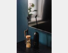 Schreibtisch mit minimalistischer Dekoration: eine hohe schwarze Vase in Form eines Gesichts, ein Glasbehälter mit getrockneten Blumen und eine schlanke Holzfigur. Im Hintergrund ist ein Teil eines Flachbildschirms und das verschwommene Bild einer Topfpflanze vor einem Fenster erkennbar.