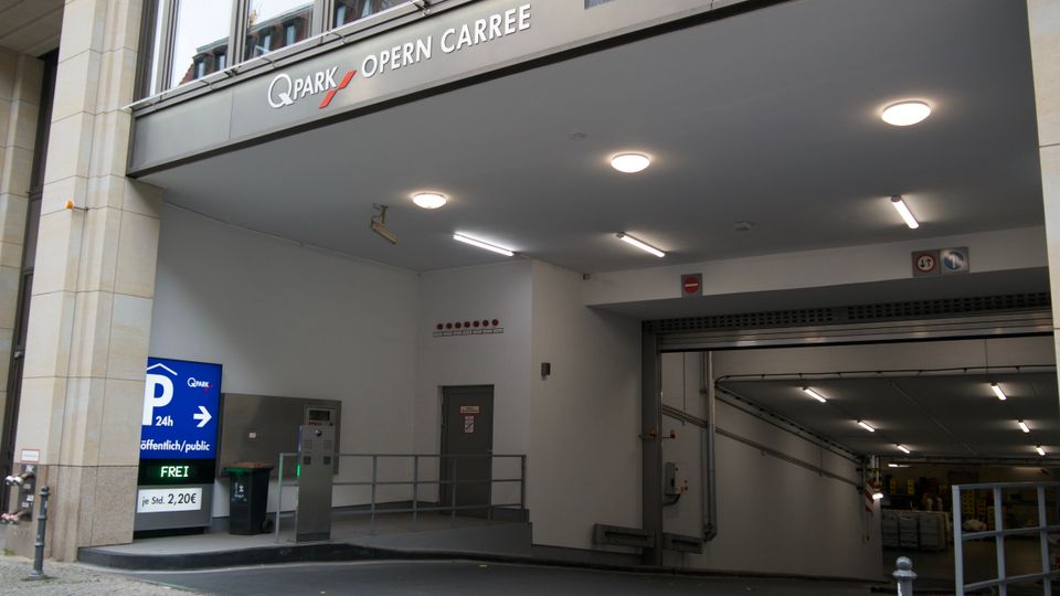 Einfahrt zu einem öffentlichen Parkhaus mit der Beschriftung „Q-Park Opern Carree“. Die Einfahrt verfügt über moderne Beleuchtung, eine Schranke, ein Hinweisschild mit Parkgebühren und Richtungsangabe sowie eine Rampe, die in den Parkplatz führt.