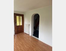 Leerer Eingangsraum mit hellem Laminatboden und weißen Wänden, links eine Holz-Eingangstür mit Glasfüllung. Daneben ein Heizkörper und ein Rundbogen-Durchgang in einen Flur mit dunklem Boden, von dem eine Holztreppe nach oben führt.