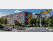 Wohnblock in einer städtischen Umgebung mit grauer Fassade, dessen Giebelseite ein großes, farbenfrohes Wandgemälde mit Blumenmotiven zeigt. Im Vordergrund befindet sich ein Kreisverkehr mit Begrünung, Verkehrsschildern und einem Gehweg, auf dem eine Person mit Rollator zu sehen ist.