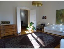 Wohnzimmer mit Parkettboden und hellem Wandanstrich, einem großen orientalischen Teppich in der Mitte des Raumes, einer weißen Couch, einem Holzkonsolentisch und Hauspflanzen. Im Hintergrund steht eine geöffnete Doppeltür, die zu einem weiteren Raum führt.