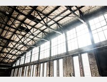 Innenansicht einer Industriehalle mit hohen Decken und sichtbaren Stahlträgern. Große, rechteckige Fenster lassen viel Tageslicht in den Raum, und die Wände bestehen aus rohem, gealtertem Mauerwerk.