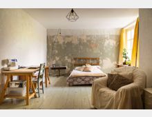 Schlafzimmer mit rustikalem Charme, das aus einem Bett mit Holzkopfteil, einem Schreibtisch mit einer Nähmaschine und einem gemütlichen Sessel besteht. Die Wände sind teils verputzt, teils abblätternd, und ein großes Fenster mit gelben Vorhängen bringt natürliches Licht in den Raum.