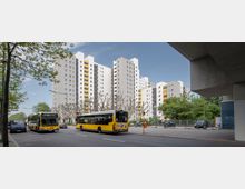 Mehrstöckige Wohnblöcke mit weißer Fassade und gelben Balkonen stehen im Hintergrund, davor eine Straße mit parkenden Autos, zwei gelben Stadtbussen und kahlen Bäumen. Rechts und links sind Gehwege und Grünflächen sichtbar.