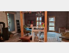 Wohnraum mit rustikalem und modernem Stil, ausgestattet mit einem weißen Regal, hölzernen Balken, einem Kronleuchter und einem Kaminofen. Die Wände sind unverputzt, der Boden besteht aus Steinfliesen, und große Fenster sowie eine Glastür bieten Blick ins Freie.