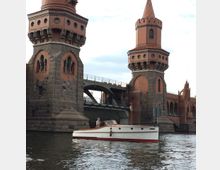 Ziegelrote Oberbaumbrücke in Berlin mit zwei Türmen und Steinfundamenten. Im Vordergrund ein weißes Motorboot auf der Spree.