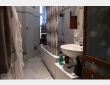 Badezimmer mit weiß gefliesten Wänden und einem weißen Waschbecken, daneben eine Badewanne mit einer geometrisch gemusterten Duschvorhang. Im Hintergrund steht ein Regal mit Handtüchern vor einem Fenster.