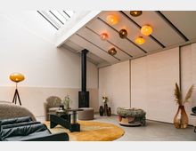 Modernes Wohnzimmer mit hellem, minimalistischem Design, einer großen Glasdecke, einem schwarzen Kamin und cremefarbenen Wänden. Der Raum ist mit gemütlichen Möbeln, einer orange-gelben Teppichfläche und dekorativen Elementen wie Vasen und einer Stehlampe ausgestattet.