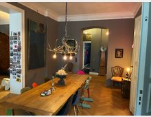 Esszimmer mit langem Holztisch und unterschiedlich farbigen Stühlen, darüber eine hängende Geweih-Lampe mit sichtbaren Glühbirnen. Graue Wände mit großen Bildern, Stuckdecke und Fischgrätparkett; im Hintergrund ein Rundbogen-Durchgang und eine Standuhr.