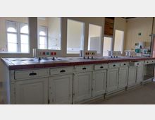 Ein heller Raum mit einer langen Arbeitsfläche aus roten Fliesen, darüber Steckdosenleisten auf Metallstützen. Darunter befinden sich weiße, abgenutzte Schränke mit mehreren Schubladen und Türen; im Hintergrund sind hohe Fenster mit Rundbögen und helle Wände zu sehen.