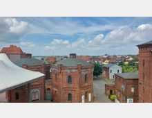 Blick auf historische Backsteingebäude mit flachen Kuppeldächern im Vordergrund, umgeben von weiteren Gebäuden und einer Kleinstadtlandschaft im Hintergrund. Der Himmel ist teilweise bewölkt, und im Hintergrund sind grüne Bäume sowie vereinzelte moderne Bauwerke zu sehen.