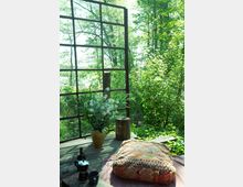 Ein rustikaler Sitzplatz im Freien mit einem großen Metallrahmen, der wie ein Fenster wirkt, umgeben von dichtem, grünem Wald. Auf einem Holztisch stehen eine gelbe Vase mit weißen Blumen, ein kleiner Holzstamm als Hocker und eine orientalisch gemusterte Sitzauflage, daneben eine Kaffeekanne und Tasse.