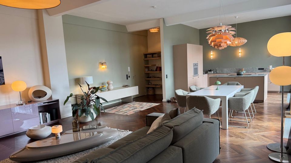 Open-plan modern living and dining area with a sectional sofa, low coffee table, and a long white dining table leading into a minimalist kitchen. Sage-green walls, herringbone wood floors, and multiple pendant and wall lights define the space, with built-in shelving and sparse decor.