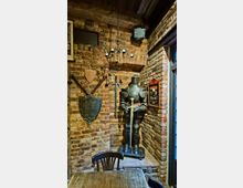 Innenraum mit rustikalen Backsteinwänden und mittelalterlicher Dekoration: eine Ritterrüstung mit Schwert in einer Ecke, daneben Schilde und gekreuzte Waffen an der Wand. Über der Ecke hängt ein schmiedeeiserner Kronleuchter; rechts befindet sich ein Fenster und ein Wandkasten-Schild.