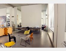 Ein offenes Büro oder Atelier mit hohen Decken und weißen Wänden, ausgestattet mit verschiedenen Stühlen, einem großen Arbeitstisch und Regalen im Hintergrund. Rechts sind große Fenster mit Jalousien, die viel Tageslicht hereinlassen, und links befinden sich Holzkisten auf Rollen.