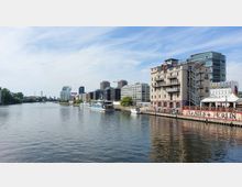 Stadtansicht an einem Fluss mit modernen Bürogebäuden, einem restaurierten Industriegebäude und Anlegestellen. Im Vordergrund ist ein Boot vertäut, und eine Uferpromenade mit Grafitti und Sitzbereichen ist sichtbar.