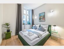 Helles, modern eingerichtetes Schlafzimmer mit grünem Polsterbett, weißer Bettwäsche und zwei Nachttischlampen. Große Fenster mit grauen Vorhängen, Holzfußboden sowie ein „Berlin“-Wandbild und ein kleiner Beistelltisch mit Pflanze ergänzen den Raum.