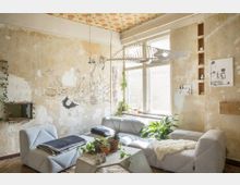 Wohnzimmer mit rustikalem Charme, gekennzeichnet durch eine Wand mit abgeblättertem Putz, einer Wandzeichnung und kleinen Regalen. Moderne graue Sitzmöbel, ein Couchtisch mit Pflanzen und große Fenster sorgen für eine helle Atmosphäre.