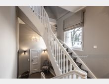 Heller Treppenhausbereich mit weißem Geländer, einer geschwungenen Treppe und großen Fenstern, die Tageslicht hereinlassen. Eine kleine Tür, eine gemusterte Sitzgelegenheit und dezente Wandlampen ergänzen den Raum.