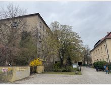 Eine Schulanlage mit zwei Gebäuden: links ein mehrstöckiges, älteres Gebäude aus Putzfassade, teilweise bewachsen, und rechts ein helles, einfaches Gebäude mit Walmdach. Im Vordergrund führt ein gepflasterter Weg durch eine kleine Grünanlage mit Bäumen und Sträuchern.