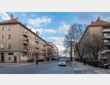 Straßenszene in einem Wohnviertel mit mehrstöckigen, hellbraunen Wohngebäuden auf beiden Seiten einer gepflasterten Straße. Am Straßenrand stehen geparkte Autos sowie einige Bäume, teils noch junge, gestützte Bäume; Fußgänger und ein Radfahrer sind ebenfalls zu sehen.