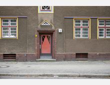 Fassade eines Wohngebäudes mit einer roten Eingangstür und Fenster mit gelben Rahmen. Die Wand besteht aus braunem Putz, die Tür- und Fensterumrandungen sind mit roten Backsteinen gestaltet.