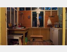 Ein einfacher Küchenraum mit Holzwänden, einem Fenster in der Mitte und einer Arbeitsplatte auf der linken Seite, auf der Küchenutensilien und ein tragbarer Herd stehen. Rechts befindet sich ein kleiner Kühlschrank, daneben stehen Wasserflaschen, und auf dem Boden liegt ein bunter Teppich.