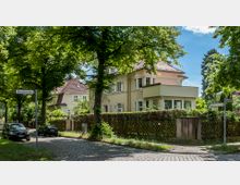 Straßenansicht eines Wohnviertels mit gepflasterten Straßen, umgeben von Bäumen. Im Vordergrund stehen zwei Autos, und im Hintergrund befinden sich Einfamilienhäuser mit roten Ziegeldächern und hellen Fassaden, eingefasst von Gärten und Holzzäunen.