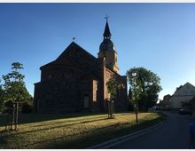 Historische Kirche mit gelbem Turm und dunklen Steinwänden, umgeben von einer Rasenfläche und vereinzelten Bäumen, bei klarer Abenddämmerung. Eine Straße führt rechts an der Kirche vorbei, gesäumt von Laternen und Wohnhäusern.