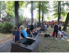 Eine Grünanlage mit großen Bäumen, in der ein Filmset eingerichtet ist. Menschen sitzen auf Stühlen und am Boden, umgeben von Filmgeräten, Kameras und Requisiten, während Kinder auf einem gemauerten Brunnenrand sitzen.