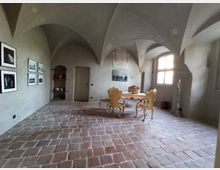 Ein historisch wirkender Raum mit Gewölbedecke, Steinfliesenboden und einem großen Fenster, das Licht hereinlässt. An einer Wand hängen Schwarz-Weiß-Fotografien, und im Raum stehen ein verzierter Tisch und zwei goldfarbene Stühle.