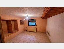 Ein leerer Kellerraum mit gefliestem Boden, orange gestrichenen Wänden und einer weißen Decke mit einfachem Lichtanschluss. Ein kleines Fenster mit Rollladen, ein Heizkörper und Holzelemente, darunter Regale und eine Deckenkonstruktion, sind sichtbar.