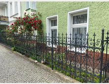 Gründerzeitliches Wohnhaus mit hellgrüner, verputzter Fassade und weißen Fenstern; davor ein schwarzer, reich verzierter Metallzaun und ein Busch mit roten Rosen. Der Boden besteht aus einem gepflasterten Gehweg.