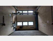 Ein leerer, kargen Raum mit betonierten Wänden, mehreren kleinen Fenstern im oberen und mittleren Bereich und freiliegenden Rohren an der linken Wand. An der linken Wand befindet sich außerdem ein kleines Waschbecken, und von der Decke hängen Metallträger.