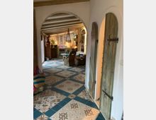 Ein Wohnraum im mediterranen Stil mit verzierten Bodenfliesen in Blau- und Beigetönen, Holzbalkendecke und bogenförmigen Türen. Im Hintergrund sind rustikale Holzmöbel, Pflanzen, geflochtene Stühle und eine dekorative Hängelampe zu sehen.