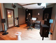 Ein modernes Tonstudio mit schallgedämmten Wänden, hölzernem Boden, einem Mischpult, mehreren Lautsprechern auf Ständern und einem Computer-Arbeitsplatz. Durch eine Glastür ist ein angrenzender Raum sichtbar.