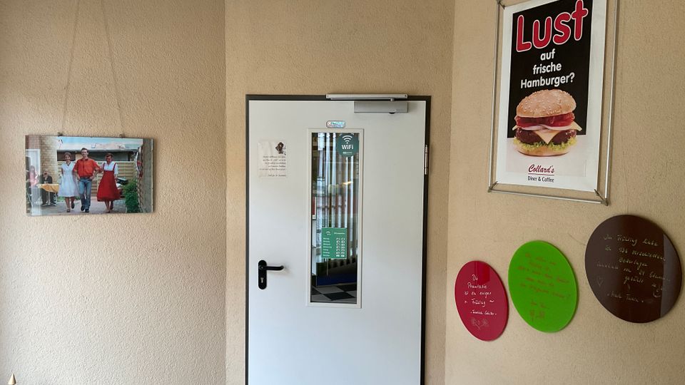 Innenbereich eines Diners oder Cafés mit beigen Wänden und einer weißen Tür mit schmalem Sichtfenster in der Mitte. An den Wänden hängen ein Burger-Werbeplakat, ein Foto sowie drei runde, farbige Tafeln mit handschriftlichen Notizen.