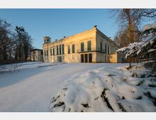 Schlossähnliches Gebäude mit hellem Putz und grünen Fensterläden, flankiert von hohen Bäumen. Der Vordergrund und die Umgebung sind mit Schnee bedeckt, einzelne Pflanzen ragen aus der Schneedecke hervor.