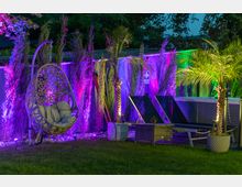 Beleuchteter Gartenbereich bei Nacht mit einer Hängesitzschaukel, zwei Sonnenliegen und einer dekorativen Anordnung von Pflanzen wie Palmen und Zypressen, die in bunten Lichtern in Lila und Grün angestrahlt werden. Im Vordergrund ist eine Rasenfläche sichtbar.