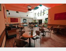 Ein moderner Innenraum eines Cafés mit Holzboden, runden Tischen und verschiedenartigen Stühlen. An der Rückwand befinden sich ein Tresen, Pflanzen und farbige Wanddekorationen, während die Decke mit orangeroten Stoffelementen gestaltet ist; an einer weißen Wand sind Schattenbilder von Personen beim Sport zu sehen.