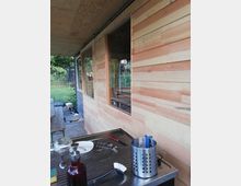 Holzwand eines modernen Anbaus mit großen Fenstern, davor eine Arbeitsfläche mit Küchenutensilien wie Besteck, Spülbürste und Seifenspender; im Hintergrund ein Garten mit Bäumen und einem Trampolin.